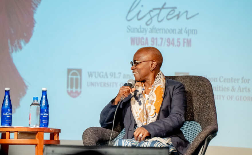 A woman wearing glasses, a dark jacket and a patterned scarf sits onstage holding a microphone during a speaking event.