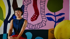 a woman sits with her hands folded on her knees with a tight smile to the camera