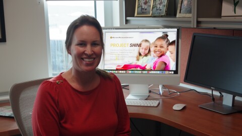 A woman in a red blouse sits in a desk chair, smiling towards the camera. A computer monitor behind her displays an advertisement for Project SHINE, with a young girl in a pink dress seen smiling in the ad. 