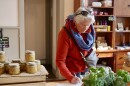 An older woman wearing a red jacket and blue scarf writes feedback on a sheet of paper on a big wooden table. Around her are jars, soaps, and bunches of basil.
