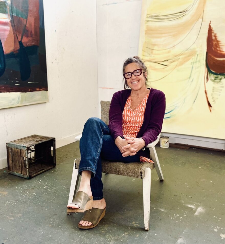 A woman sits in a chair and smiles at the camera. Some paintings can be seen in the background