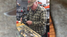 A man wearing a plaid patter shirt measuring silver knives with a table with a pile of knives in front of him.