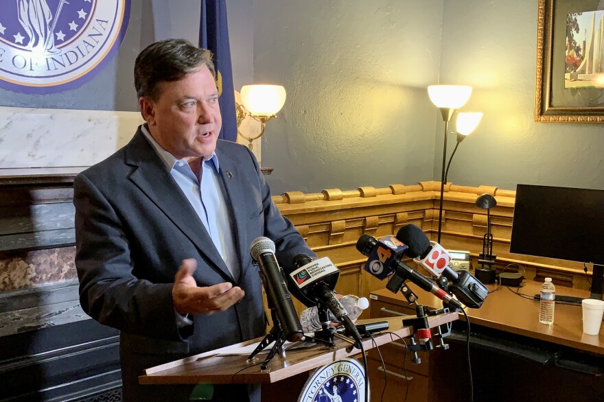 Indiana Attorney General Todd Rokita speaks to reporters in his office at the Statehouse. In front of Rokita is a lectern, on which sit several microphones. Behind him is a fireplace with the seal of the Office of the Attorney General on the wall above it.