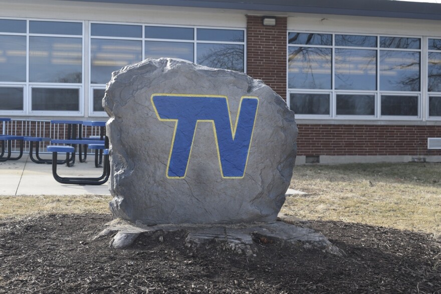 A large rock with the letters TV painted in blue with a gold outline placed next to a school building.