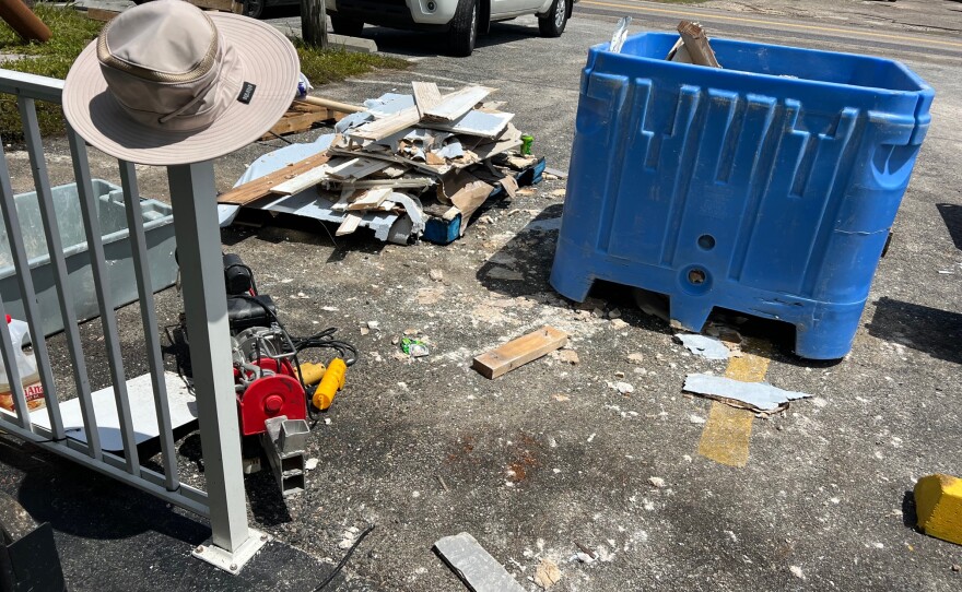 Storm debris and building materials are piled in a parking lot. A sunhat dangled from the railing.