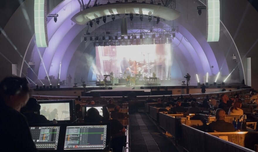 A view of the Hollywood Bowl shell and stage from behind the sound control booth. We see sound controls, some floor seats, and the stage, as well as the L-Acoustics speakers, which are in the giant white columns hanging from the Bowl's shell.