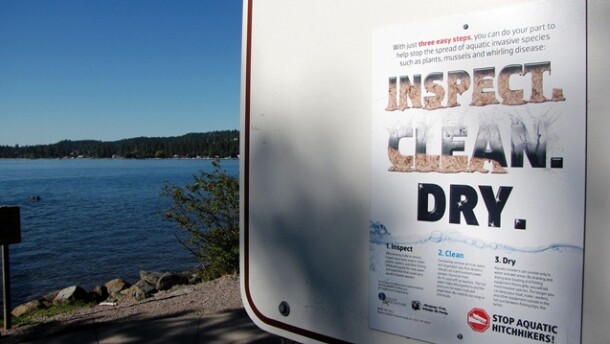 A sign in the foreground gives information about preventing the spread of aquatic invasive species. A large body of water is visible in the background.