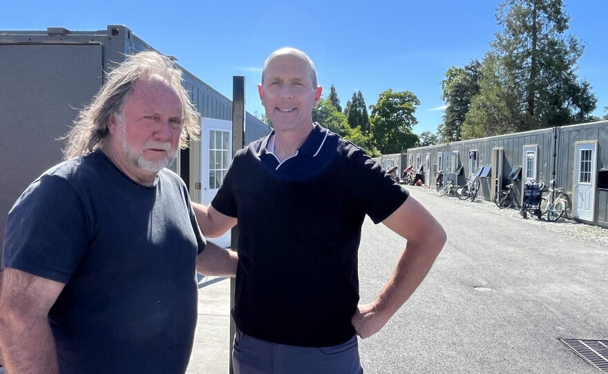 Two middle-aged men are standing next to each other outdoors. Behind them are shipping container homes. 