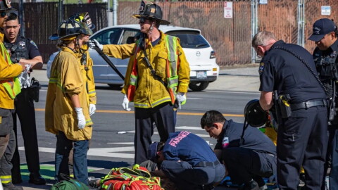 Firefighters in yellow jackets stand on the left while paramedics in blue attend to a person lying on the ground.
