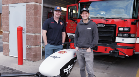 Two men stand in front of a fire engine and beside a small drone, about the size of a bicycle
