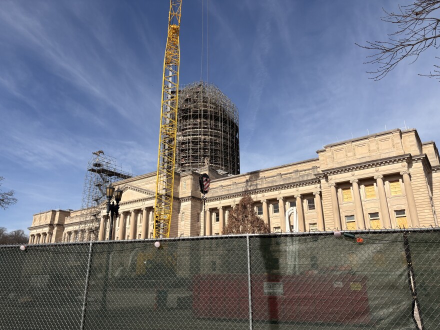 Kentucky's state Capitol building. The photo is taken from the other side of a construction fence lined with semi-translucent green fabric. Between the Capitol and the fence is a large crane which extends out of frame. The dome is covered in scaffolding.