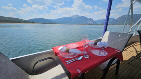 a table set for two on the deck of a boat has a red tablecloth, two beige and brown chairs. It's overlooking the water of Lake Dillon with mountains in the background
