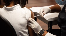 A young girl gets a COVID-19 vaccine in her right arm. She's wearing a white shirt and her back is turned. The nurse is in blue scrubs, a black watch visible on her wrist, administering the vaccine.