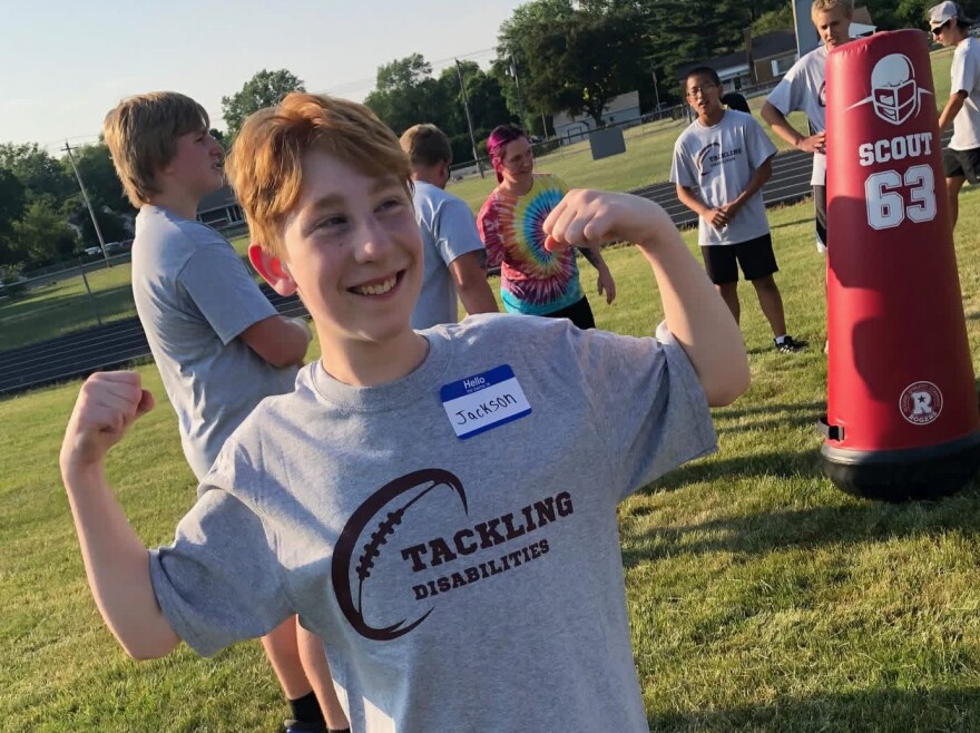 A smiling boy with light skin and red hair flexes his arms, wearing a "Tackling Disabilities" t-shirt and a name tag that says "Jackson." Other kids and a football tackling dummy are visible in the background on a grassy field.