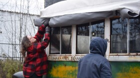 A woman in a red buffalo-checked shirt reaches up to roll up a tarp that was covering the iconic green city bus that Chris McCandless died in.