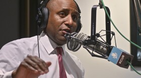A man inside a radio studio sits behind a microphone. He is talking and gesturing with hie right hand.