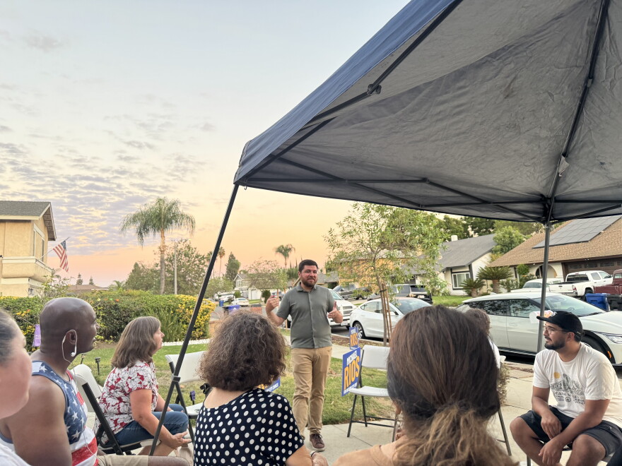 A man talks facing the camera on a driveway in a neighborhood while people watch under a tent 