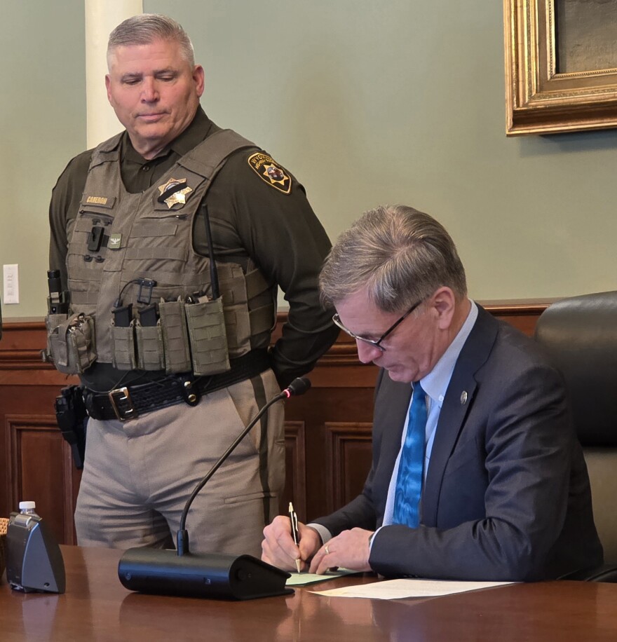 A police officer with his hands behind his back looks down at a desk where the governor is sitting and signing a bill into law.
