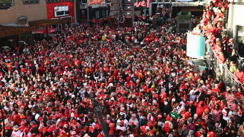 A crowd of thousands pack together inside a venue. There are many lighted signs for restaurants around and above the crowd.
