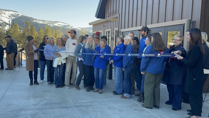 Dozens of people stand behind a blue ribbon. A lady in the middle of the crowd holds big scissors 