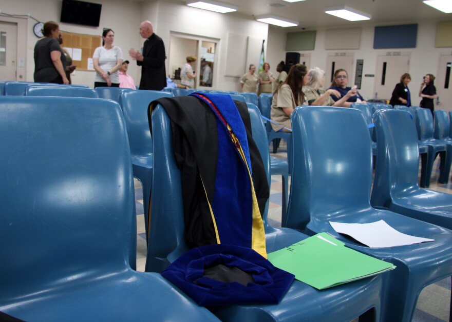 A PhD cap and gown sits on a blue plastic chair. People stand in the background talking.