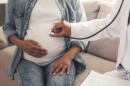 Doctor and pregnant patient, who are Black, sit on a couch. Doctor holds stethoscope to patient's chest while she rubs her belly.