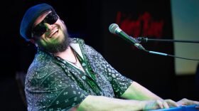A musician wearing sunglasses and a cap smiles while playing a keyboard onstage.