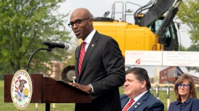 Standing in front of Gov. J.B. Pritzker and Rep. Katie Stuart, Chancellor James Minor of Southern Illinois University Edwardsville speaks behind a podium on a sunny fall day.