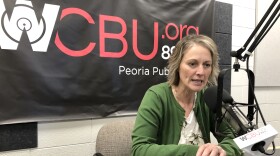 A woman wearing a green sweater speaks into a microphone as she sits at a table in front of a wall featuring a WCBU banner. 