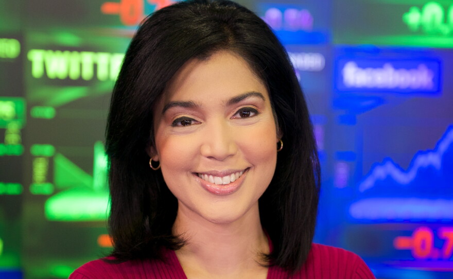 A woman with shoulder-length dark hair is wearing a red sweater and standing with her arms crossed in front of a wall of monitors showing financial graphs.