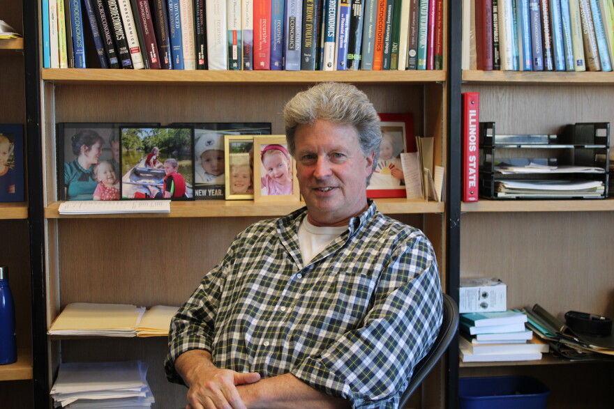 A man sits in front of a bookshelf full of textbooks and photos and smiles. 