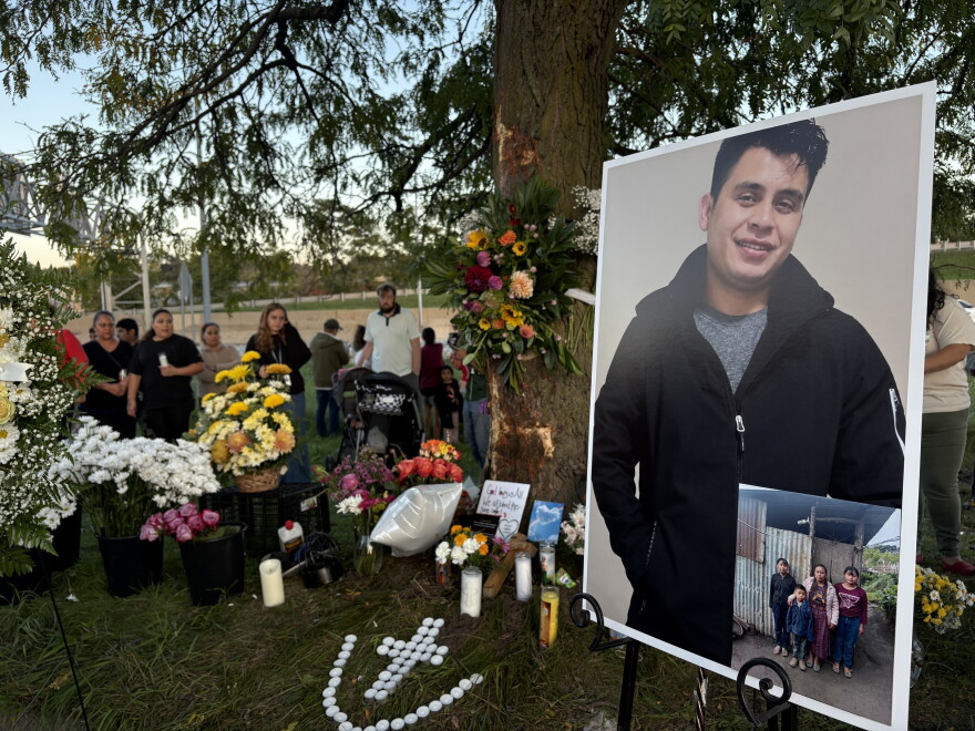 A poster-sized photo of a smiling man in a dark zip up sweater sits on an easel in front of a treet surrounded by colorful flowers. On the ground, white tea lights are arranged in the shape of a cross. 