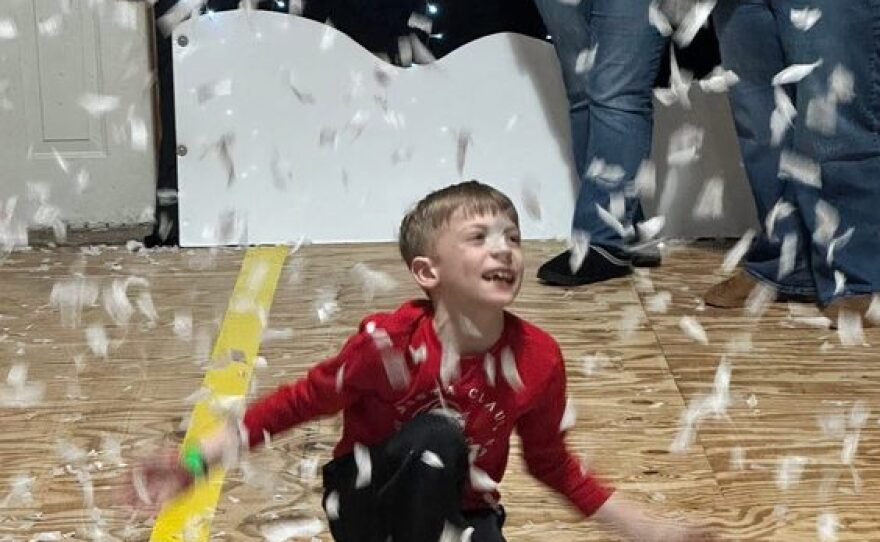 A young boy wearing a red shirt joyfully plays in an indoor area with white feathers falling around him, smiling and crouching on a wooden floor.