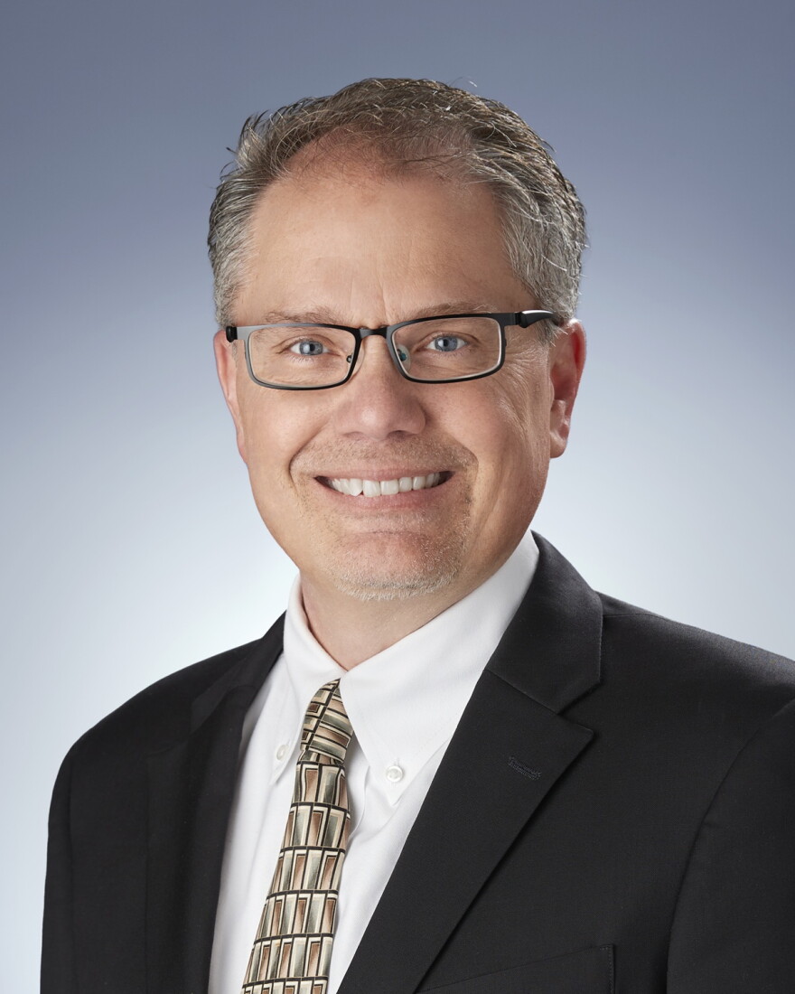 Portrait of man in a dark suit, white shirt and patterned tie and wearing glasses