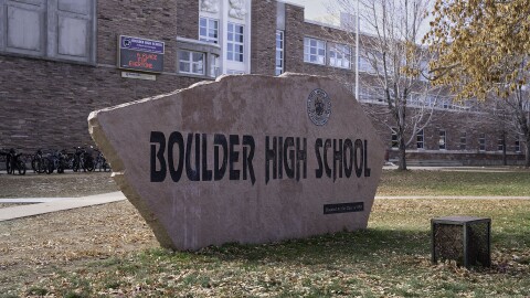 A slab of reddish grey sandstone with black embossed letters that read Boulder High School sits on a lawn in front of a brick building in the late autumn in the American West