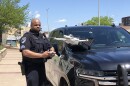 A police officer operates a white drone using a remote control, standing next to a patrol vehicle on a sunny day, with buildings and trees in the background.