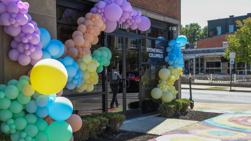 The front of a building is decorated with a garland made of balloons of various sizes and colors. In the reflection of the glass doors, a man can be seen walking. A decal on the windows reads, "Stonewall Columbus." 