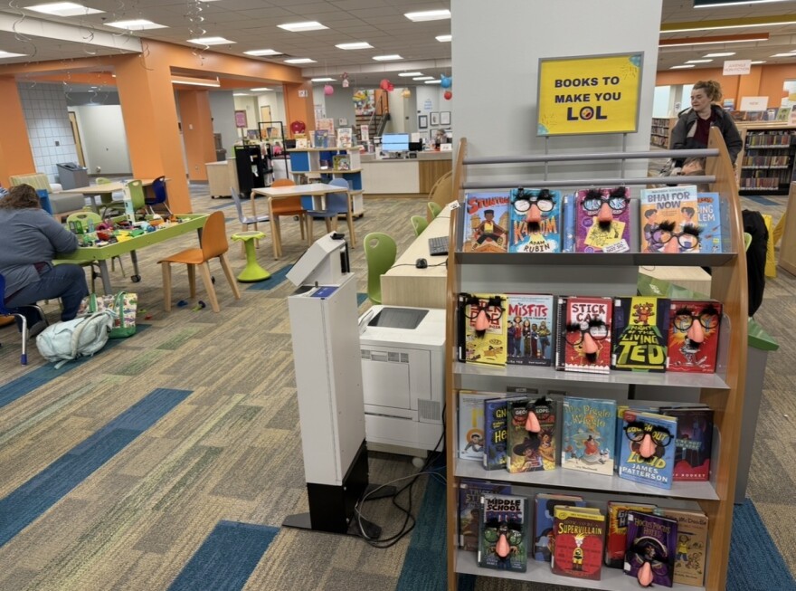 In a colorful room lined with shelves of books, one shelf of books has a sign over it that says "Books to make you LOL". On the shelf, all of the books have a pair of glasses on them with a nose and mustache "disguise" attached.