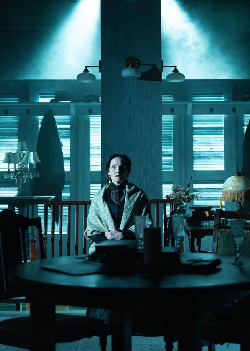 Jeanine Kane (Mrs. Helen Alving) sits at a table in a room with blue-hued lighting. 