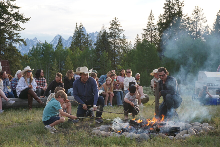 Adults and kids kneel and squat around a smoky campfire, roasting marshmallows. People sit in the background, in front of a view of rocky mountains.