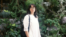 A late twenties Vietnamese person in a white shirt, dark glasses, and medium length hair stands in front of a wall of foliage. 