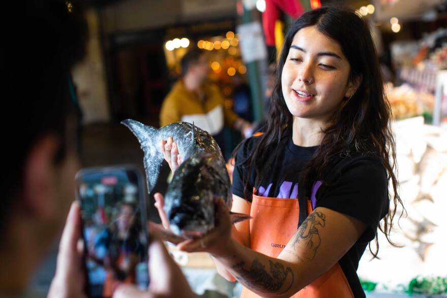  A young woman with long dark hair and tattoos holds a fish in front of a cellphone camera.