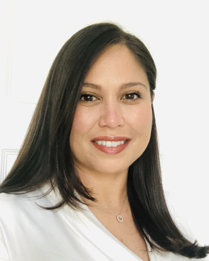 A young woman with long brown hair and brown eyes, wearing a white blouse looks at the camera.