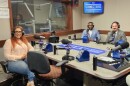 Three smiling people wearing headphones sit at a table in a radio talk studio: a woman front left has long, red, wavy hair and is wearing glasses, gold hoop earrings, a long sleeved peach top, jeans, and tan sandals; a man back left has short black hair and is wearing glasses and a white shirt under a light blue suit coat; a man back right has short black hair and a mustache and is wearing a white collared shirt and blue tie under a grey suit coat