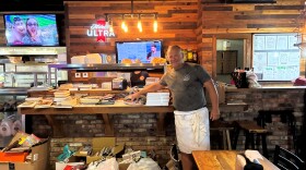 Alt text:

A man in a gray T-shirt and white apron stands smiling beside a large stack of secondhand books piled on a wooden bar counter inside a restaurant or pizzeria. More books and bagged donations fill boxes and shopping bags on the floor below. A neon Michelob Ultra sign, string lights, and exposed brick line the wall behind him, with a TV playing sports coverage and a community bulletin board visible in the background.