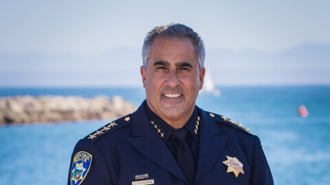 A man in a formal blue police uniform stands with his hands clasped in front of him. He's in front of a railing and behind that is the ocean with a jetty in the distance.