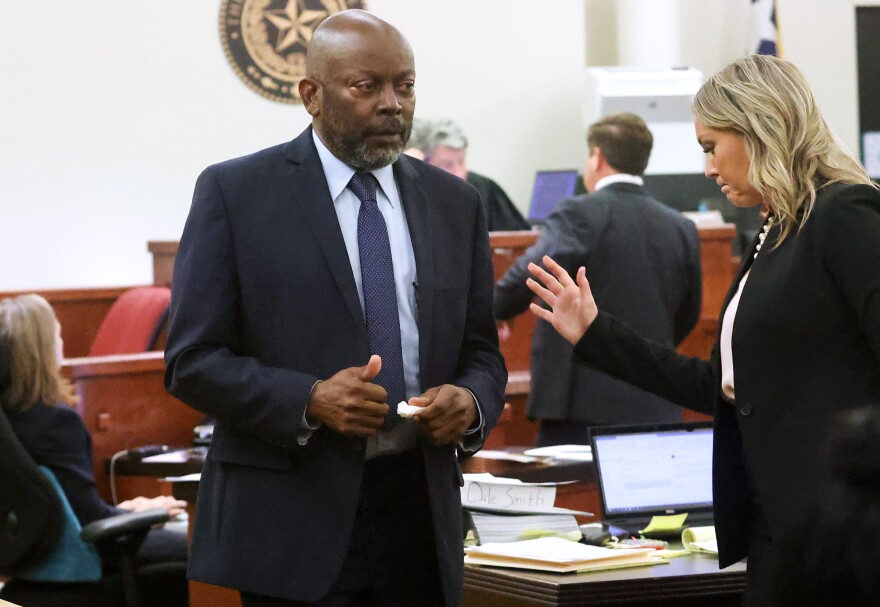 James Smith, an older Bklack man wearing a suit and tie, gives a small thumbs-up as he walks through a courtroom. Prosecutor Ashlea Deener, a white woman wearing a suit, raised her hand next to him.