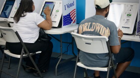 Interior photo that shows two people sitting in folding chairs with their backs to the camera. They appear to be making selections on electronic screens that are voting machines.
