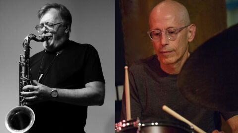 Two guys wearing black t-shirts. The one on the left playing a saxophone and the one on the right playing drums.