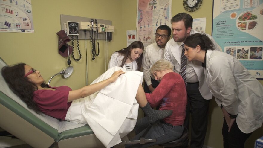 A group of medical students stand around a gynecological teaching associate who's performing a pelvic exam on a woman. 